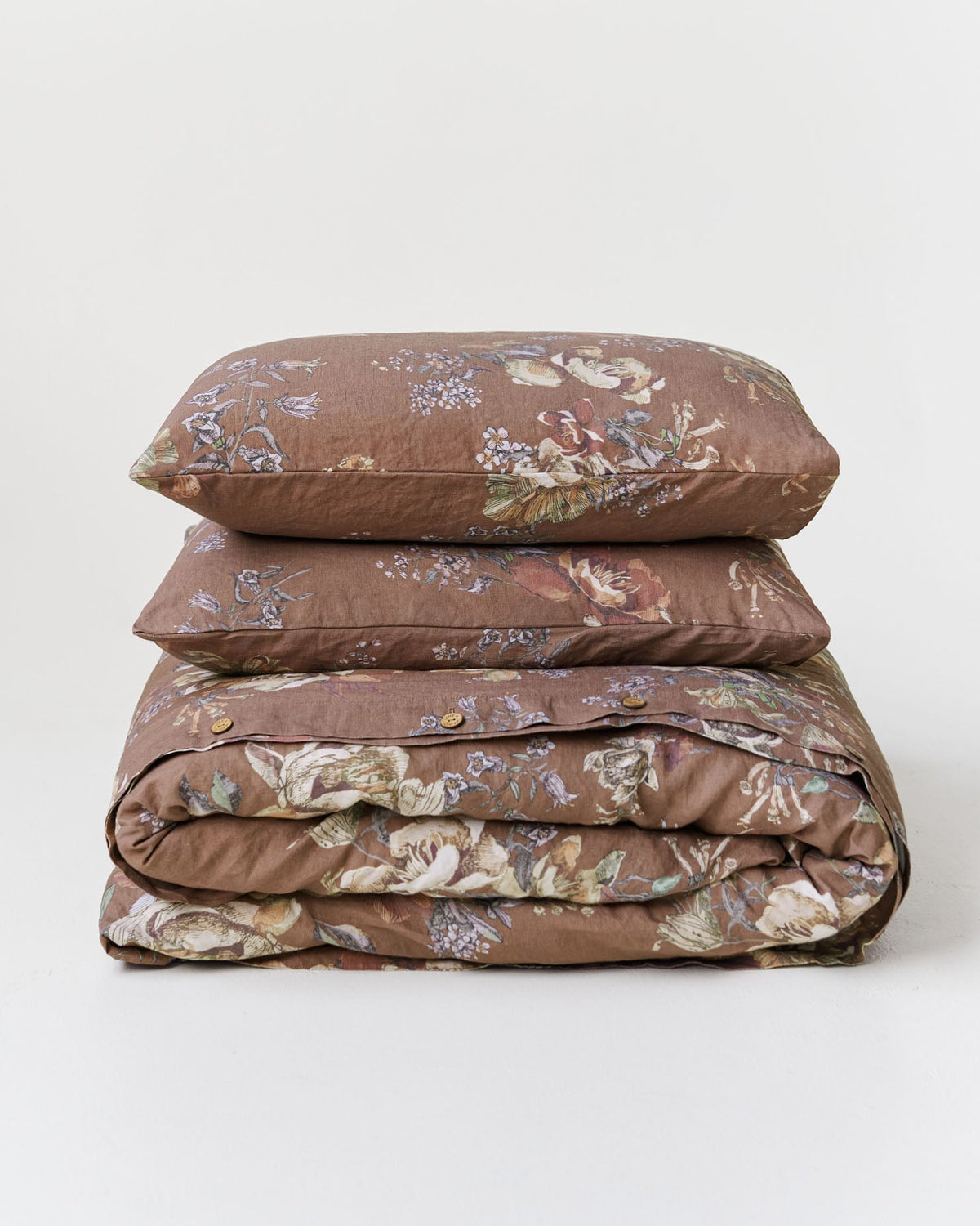 A stack of three bedding items, including two pillows and a folded duvet cover, in a brown floral pattern on a light gray background.