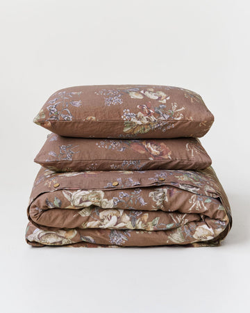 A stack of three bedding items, including two pillows and a folded duvet cover, in a brown floral pattern on a light gray background.
