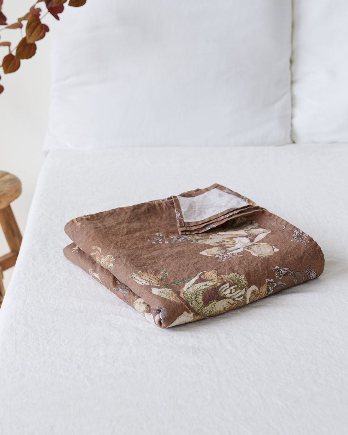 A folded brown patterned fabric item, likely a quilt or blanket, styled on a white bed next to a small wooden stool with dried leaves.